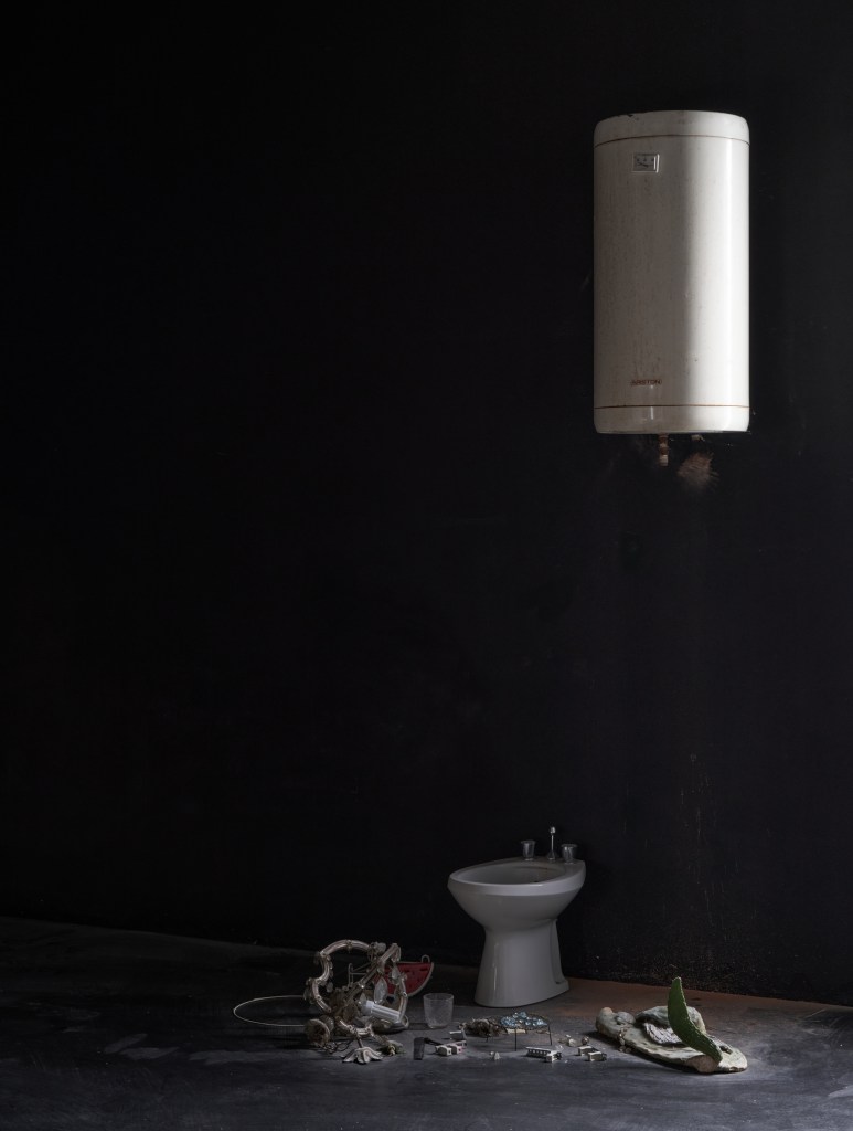 universe (2025), installation using a washing hanger from a demolished bathroom and found objects, including a water tank, toilet, and Murano glass. Dimensions variable. The work presents a poetic archaeology of abandoned domestic spaces.