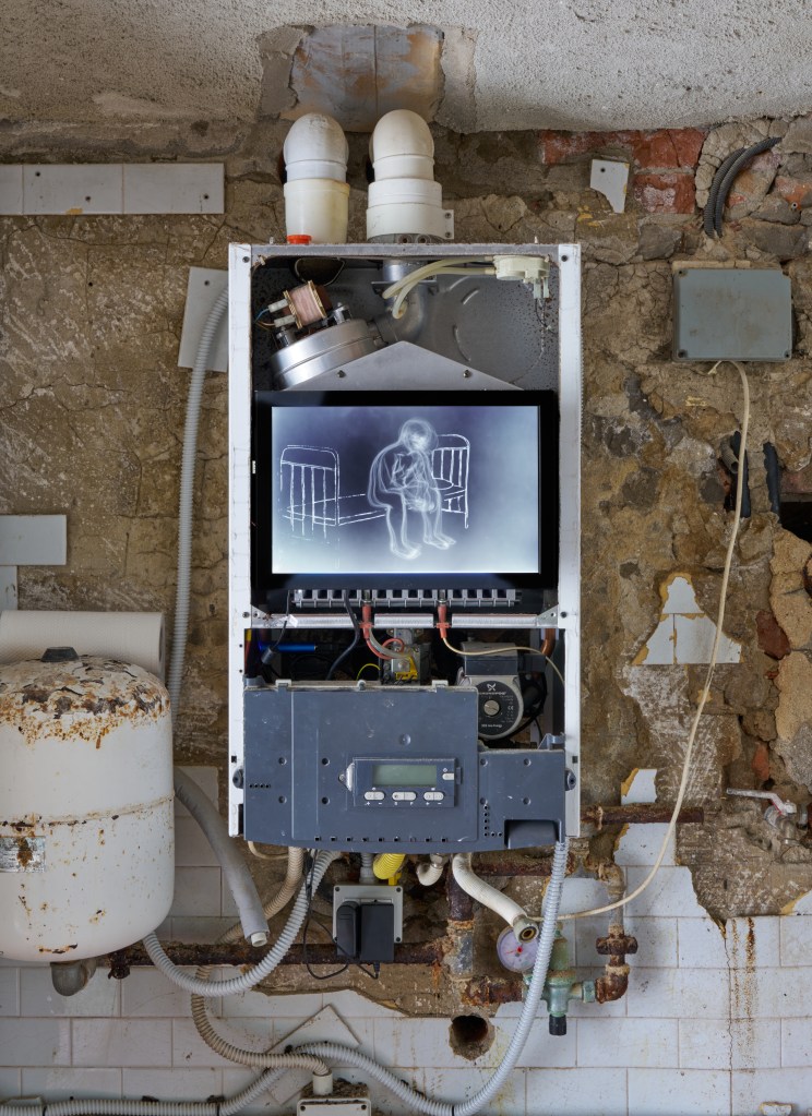 forbidden place (2025), installation composed of a room with pipes, a boiler, and a monitor. The work confronts hidden infrastructures and psychological enclosures within built environments.