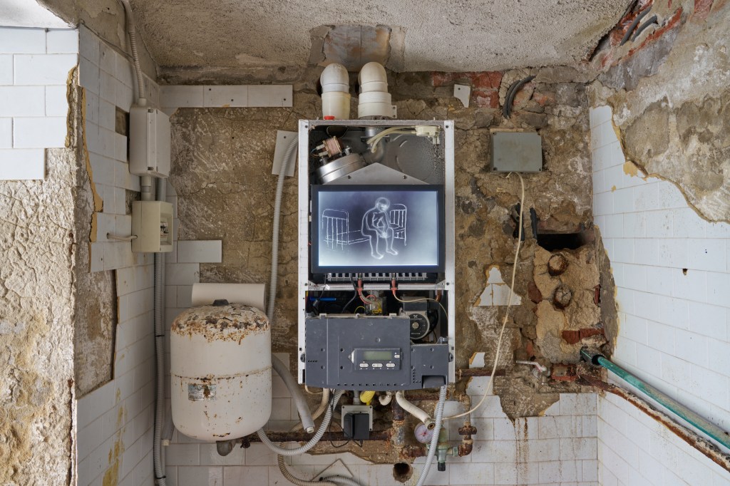 forbidden place (2025), installation composed of a room with pipes, a boiler, and a monitor. The work confronts hidden infrastructures and psychological enclosures within built environments.