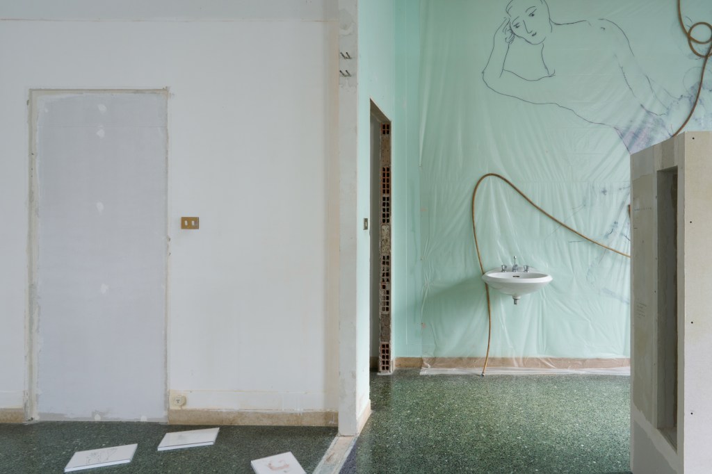 to love and devour (2025), an installation made of plastic, permanent marker, plastic tubing, and a sink, measuring 375 by 367 cm. The work evokes a fragmented, provisional environment layered with personal and industrial materials.