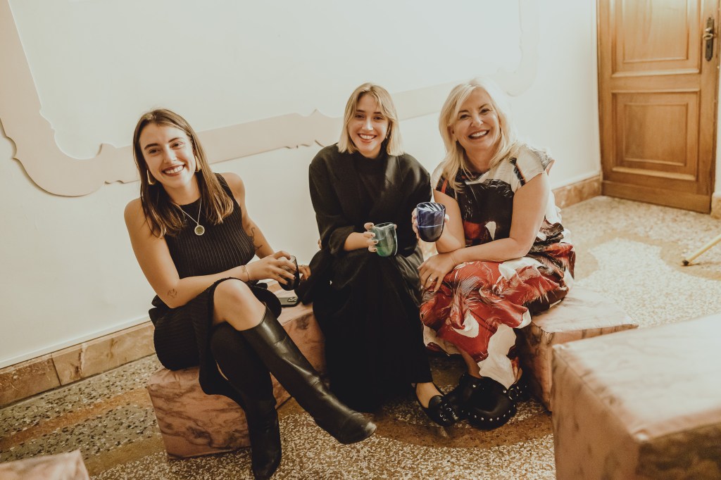 Portrait of three smiling women seated indoors on pink marble-like stools, holding colored glass cups. The scene is warm and casual. Photo by Pierre Morel, taken during Libreria delle Api by Fiorucci Colmignoli, Venice, 2025.
