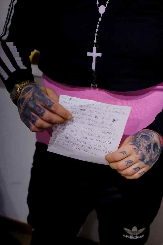 Close-up view of a person wearing a tracksuit with visible tattoos on their hands holding a handwritten note. A rosary with a cross hangs from their jacket. Photo by Francisco de Figueiredo, from Imaginary Palace, part of NERO Closer, 2024.