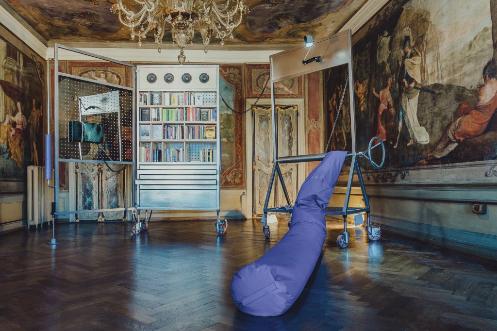 Installation view of Libreria delle Api, featuring a mobile bookshelf, hanging monitors, and a padded purple structure on wheels, set within a richly decorated frescoed room. Photo by Pierre Morel, Venice, 2025.