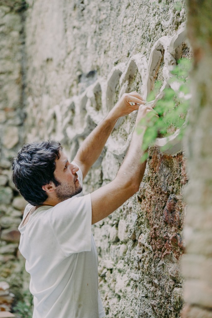 An artist working outdoors on a stone wall installation surrounded by greenery, captured during the Midsummer Daydream residency.
