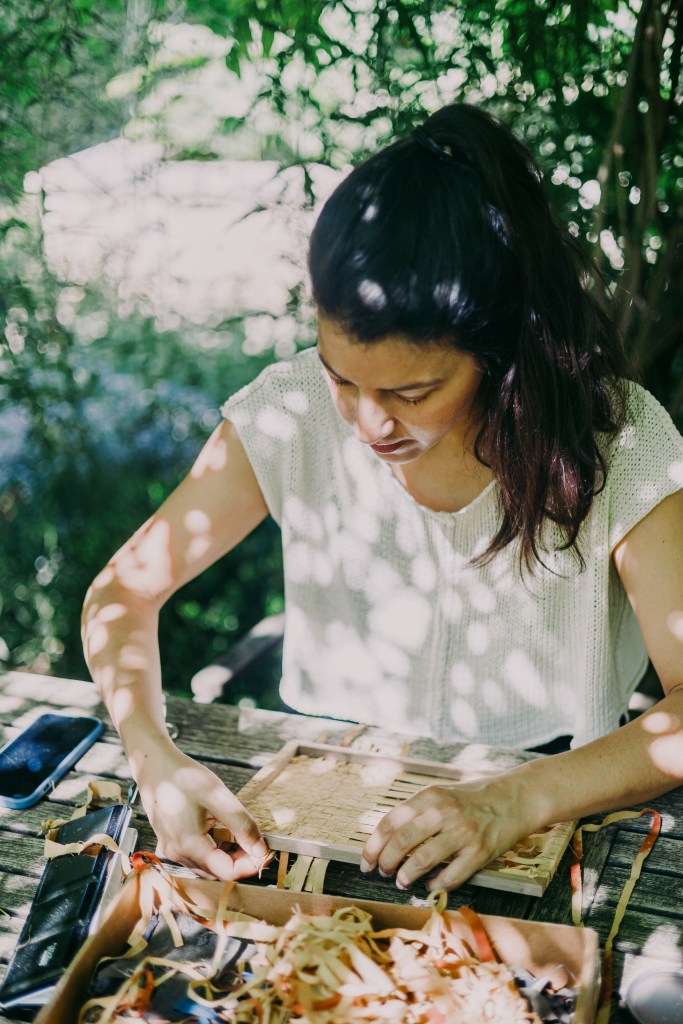 A woman weaving natural fibers at a wooden table under trees, part of Rodriguez’s process during Midsummer Daydream