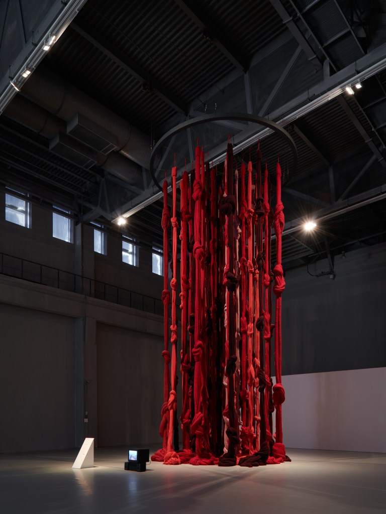 Installation view of Cecilia Vicuña’s Quipu Menstrual (Shanghai) at the 13th Shanghai Biennale, showing hanging strands of unspun wool forming a quipu-like structure combined with video elements in a large exhibition space.