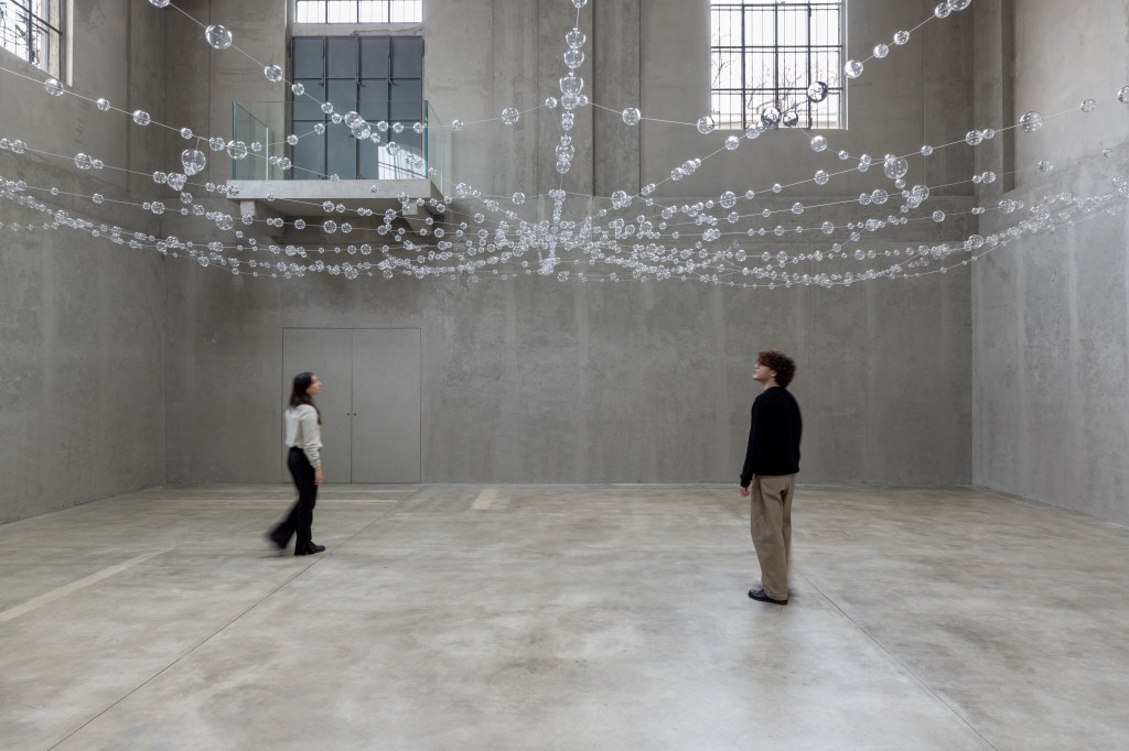 Installation view of Mona Hatoum’s Web (2026), featuring suspended clear glass spheres connected by steel cables within the exhibition space.