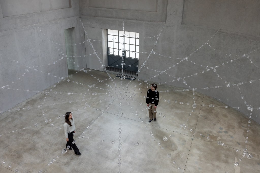 Installation view of Mona Hatoum’s Web (2026), featuring suspended clear glass spheres connected by steel cables within the exhibition space.