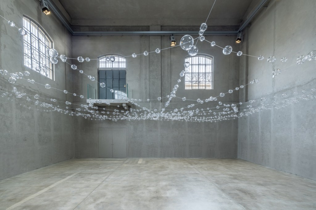 Installation view of Mona Hatoum’s Web (2026), featuring suspended clear glass spheres connected by steel cables within the exhibition space.