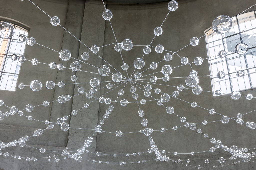 Installation view of Mona Hatoum’s Web (2026), featuring suspended clear glass spheres connected by steel cables within the exhibition space.