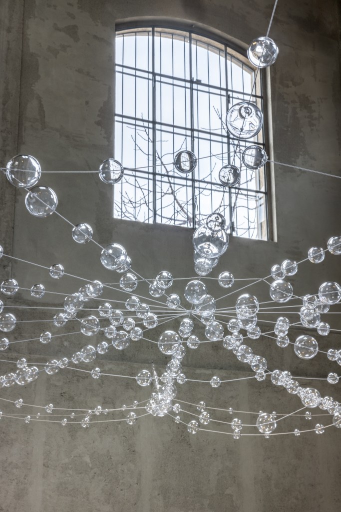 Installation view of Mona Hatoum’s Web (2026), featuring suspended clear glass spheres connected by steel cables within the exhibition space.