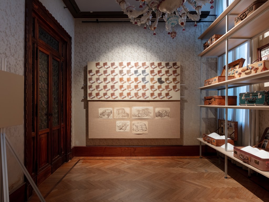 Suitcases and Shifting Sands (1974/2026) and Cases Filled with Sand (1993) by Vyacheslav Akhunov, installation view at Palazzo Franchetti. An installation of suitcases filled with or surrounded by sand, evoking migration, displacement, memory, and the passage of time