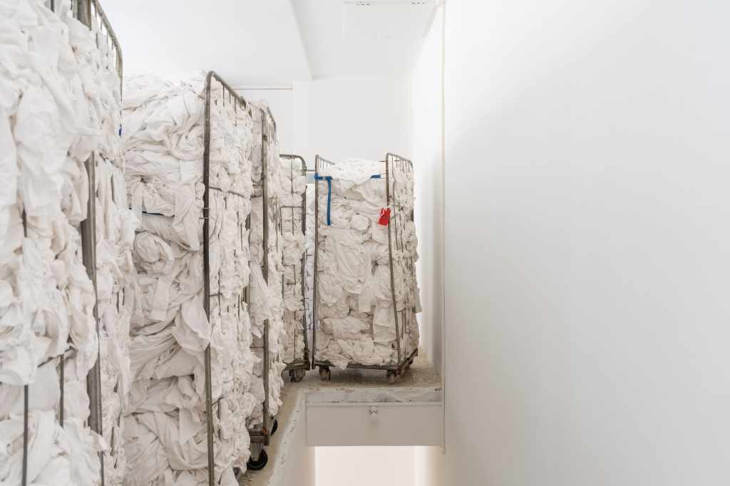 1.3 tons of decommissioned bed linen from 200 Venetian hotels: Large piles or folded stacks of white hotel bed linens fill the installation space, highlighting the scale and material presence of discarded textiles