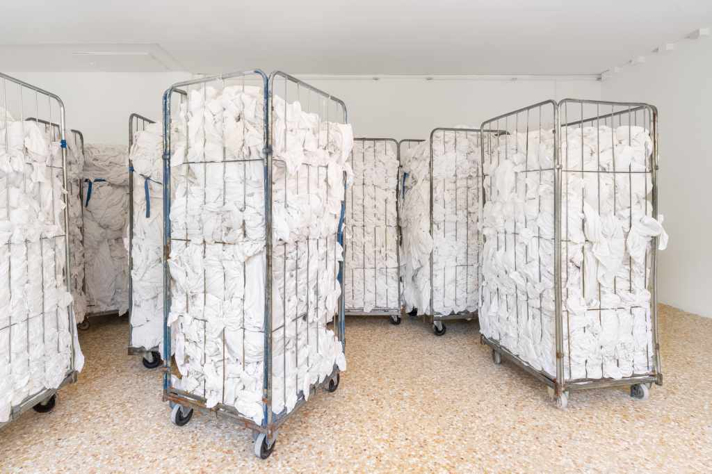 1.3 tons of decommissioned bed linen from 200 Venetian hotels: Large piles or folded stacks of white hotel bed linens fill the installation space, highlighting the scale and material presence of discarded textiles