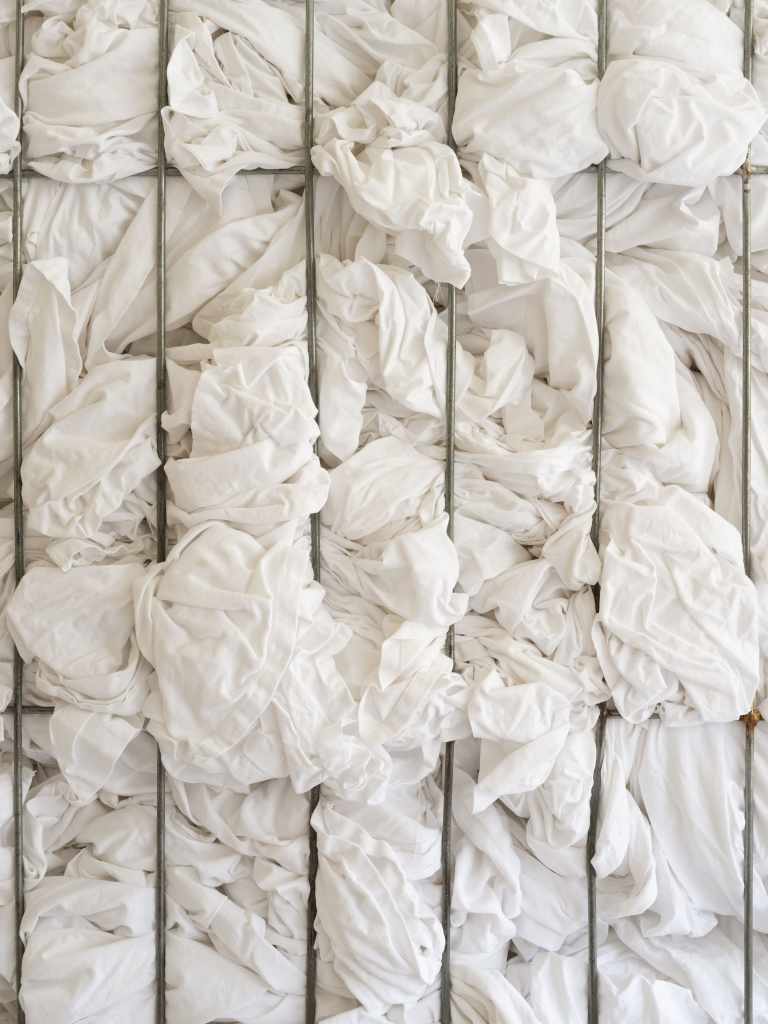 1.3 tons of decommissioned bed linen from 200 Venetian hotels: Large piles or folded stacks of white hotel bed linens fill the installation space, highlighting the scale and material presence of discarded textiles